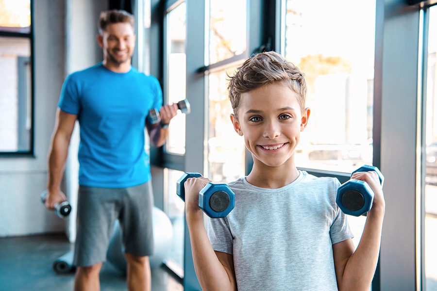 Employee Benefits - Selective Focus of Young Son and His Father with Dumbbells in Exercise Room